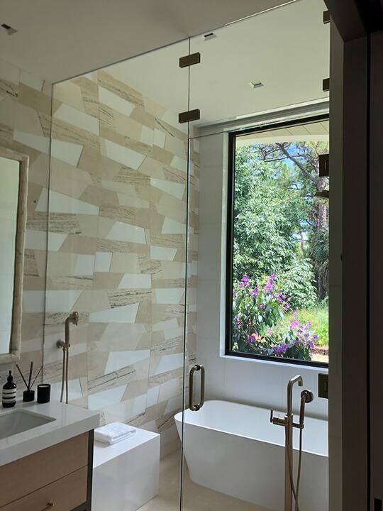 Floor-to-ceiling frameless shower glass beside a garden window and soaking tub
